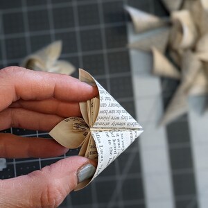 May include: A paper flower made from the pages of a book. The flower is folded into a star shape and has a light brown color. The flower is being held in a hand.