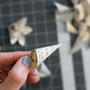 May include: A hand holding a small paper cone made from a book page. The cone is folded into a point and the text on the page is visible. The text reads "it, my" and "so odd a".