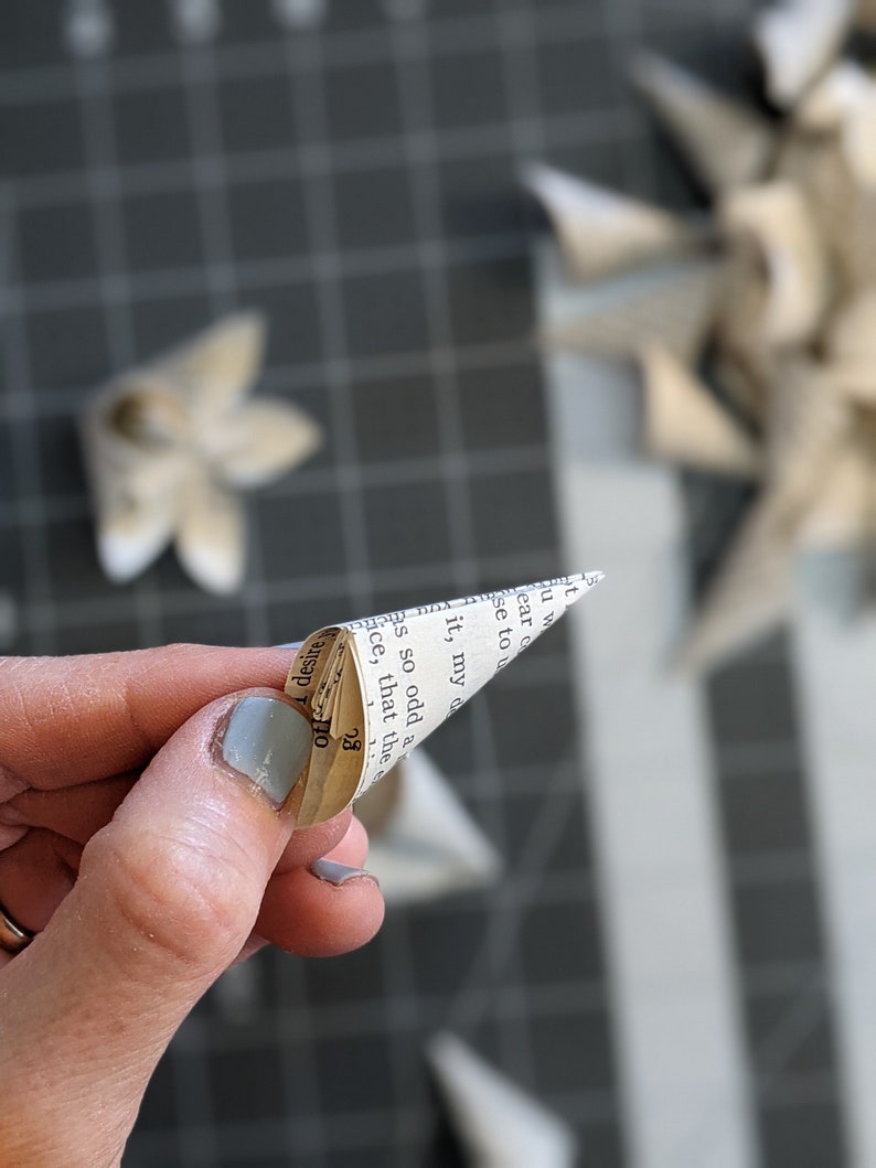May include: A hand holding a small paper cone made from a book page. The cone is folded into a point and the text on the page is visible. The text reads "it, my" and "so odd a".