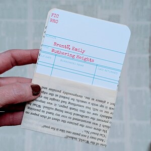 May include: A white library card with the title "Wuthering Heights" by Emily Bront&euml;. The card is held with a piece of paper with printed text.