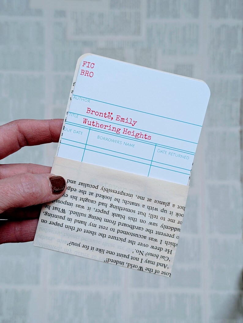 May include: A white library card with the title "Wuthering Heights" by Emily Bront&euml;. The card is held with a piece of paper with printed text.