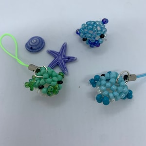 Könnte beinhalten: Drei Perlen-Meeresschildkröten-Anhänger mit grünen, blauen und hellblauen Perlen. Die Anhänger sind an einer Schnur mit einem Karabinerhaken befestigt. Die Anhänger sind von einem weißen Seestern und einer blauen Muschel umgeben.