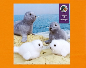 Op de afbeelding: Vier vilten dieren speelgoed, twee grijze zeehonden en twee witte zeehonden, zijn gerangschikt op een zandstrand. De zeehonden kijken allemaal naar de camera. De achtergrond is een blauwe lucht en zee. De afbeelding bevat de tekst "Vrolijke Vilt Vriendjes" in paars.