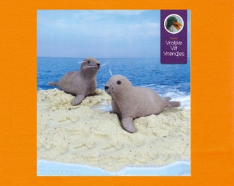 Op de afbeelding: Twee grijze vilten zeehondjes op een zandstrand met blauw water op de achtergrond. De tekst "Vrolijke Vilt Vriendjes" staat op een paars label met een groene eend illustratie.