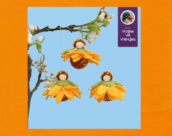 Op de afbeelding: Drie vilten ornamenten in de vorm van zonnebloemen met een vilten poppenhoofd in het midden. De ornamenten hangen aan een tak met witte bloesems. De tekst op het paarse label luidt "Atelier Vrolijke Vilt Vriendjes".