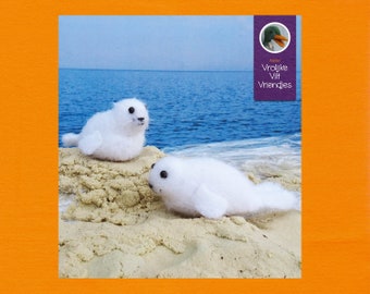 Op de afbeelding: Twee witte vilten zeehondjes op een zandstrand met blauw water op de achtergrond. De tekst "Atelier Vrolijke Vilt Vriendjes" staat op een paars label met een groene vilten eend.