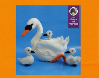Op de afbeelding: Een witte vilt zwaan met drie kleinere vilt zwanen, waarvan er twee in de grotere zwaan zitten. De grotere zwaan heeft een lange oranje snavel en zwarte ogen. De kleinere zwanen hebben zwarte ogen en oranje snavels. De achtergrond is lichtblauw.
