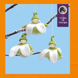 May include: Three felt snowdrop ornaments with green felt hats and faces. The ornaments are hanging from a branch with snow on it. The text on the label reads "Vrolijke Vilt Vriendjes".