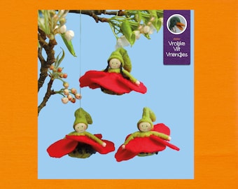 Op de afbeelding: Drie vilten poppen die aan een tak hangen. Elke pop draagt een groene hoed en een rode viltrok. De poppen hangen aan een touwtje en zijn omringd door witte bloemen.