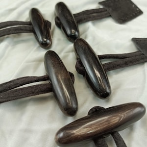 Könnte beinhalten: Fünf dunkelbraune Holz-Toggle-Knöpfe mit schwarzen Schnüren. Die Knöpfe sind oval geformt und haben eine glatte, polierte Oberfläche.