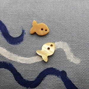 Può includere: Due piccoli bottoni di legno a forma di pesce di colore beige con due fori ciascuno. I bottoni sono su uno sfondo di tessuto grigio testurizzato.