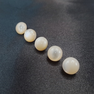 Peut inclure: Cinq boutons nacrés blancs disposés en rangée sur un fond de tissu noir.