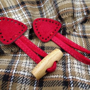 Könnte beinhalten: Zwei rote Lederknöpfe mit einem Holzknopf. Die Knöpfe sind an einem karierten Stoff befestigt.
