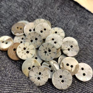 Puede incluir: Un conjunto de botones en tonos crema, beige y marrón. Algunos botones presentan un diseño floral, mientras que otros son lisos. Los botones, con dos o cuatro agujeros, están dispuestos sobre una tela azul oscuro texturizada.