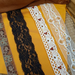 May include: Four different types of lace trim, including black, white, and brown, are displayed on a yellow fabric background. The lace trims are all different widths and have different patterns.