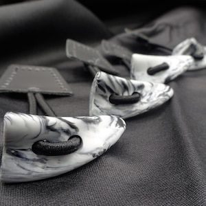 May include: Five black and white marbled toggle buttons with black elastic loops. The buttons are arranged in a row on a black fabric background.