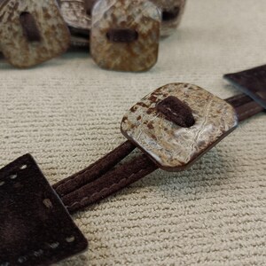 May include: A pair of brown leather toggles with a square, marbled, beige button. The toggles are connected by a brown suede strap.