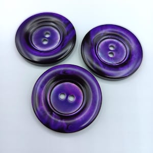 Peut inclure: Trois boutons à deux trous, de couleur violet et noir avec un motif en spirale.