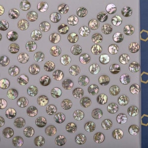 Op de afbeelding: Een verzameling van iriserende parelmoerknopen in verschillende tinten bruin, groen en goud. De knopen zijn willekeurig gerangschikt op een lichtgrijze achtergrond.