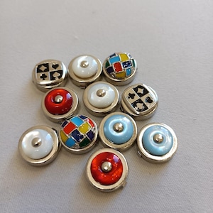 Pode incluir: Uma coleção de botões decorativos com bordas de metal prateado. Os botões apresentam vários designs, incluindo naipes de cartas de baralho, padrões geométricos coloridos e cores sólidas como vermelho e azul claro. Os botões estão dispostos sobre uma superfície cinza claro.