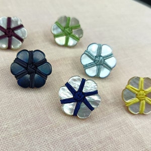 Op de afbeelding: Zes decoratieve bloemvormige knopen met een parelmoerachtige afwerking en kleurrijke touwaccenten. De knopen zijn in tinten paars, groen, blauw, lichtblauw en geel.
