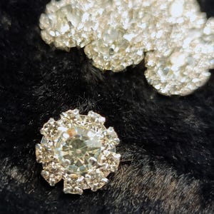 May include: A silver-toned, decorative button with a clear, faceted crystal in the center. The button is surrounded by smaller, clear crystals.
