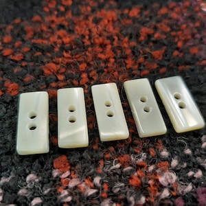 Op de afbeelding: Vijf witte rechthoekige knopen met elk twee gaten. De knopen zijn gemaakt van parelmoerachtig materiaal en hebben een gladde, gepolijste afwerking.