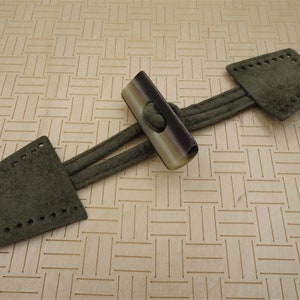 May include: A green suede toggle closure with a brown and white striped toggle button. The toggle is attached to two green suede straps.