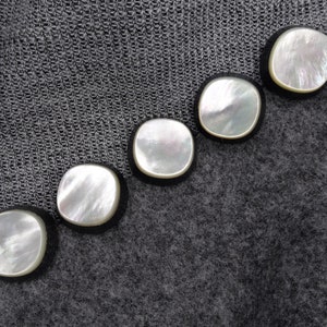 May include: Five round, white, mother-of-pearl buttons with black trim, arranged in a diagonal line on a gray fabric background.