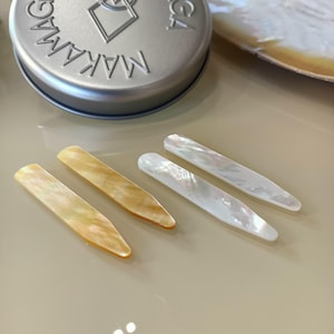 Könnte beinhalten: Vier schillernde Hemdkragenstäbchen aus Perlmutt in Gold- und Weißtönen. Eine silberne Dose mit dem Wort "MAKAMA" im Hintergrund. Die Kragenstäbchen liegen auf einer hellen Oberfläche.