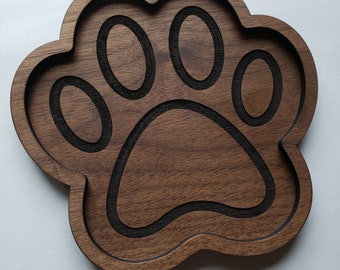 May include: A wooden paw print-shaped tray with a dark brown finish. The tray features a carved paw print design with a darker, contrasting interior. The edges are rounded, and the tray is set against a white background.