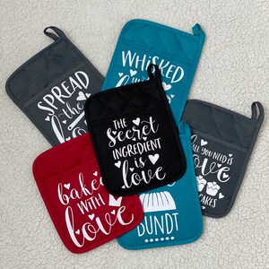 Puede incluir: Conjunto de seis manoplas para horno con diferentes lemas. Las manoplas son de varios colores, incluyendo negro, rojo, gris, azul verdoso y turquesa. Los lemas incluyen "Spread the Love", "The Secret Ingredient is Love", "All You Need is Love", "Baked with Love", "Bundt" y "Whisked with Love".