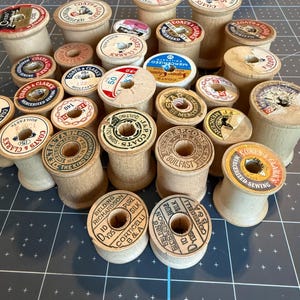 May include: A collection of vintage wooden sewing thread spools with various paper labels on the top ends. The spools are light brown in color and feature printed brand names such as Coats & Clark, J. & P. Coats, and Belding Richardson.