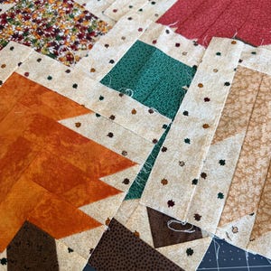 May include: A collection of fabric quilt blocks featuring tree shapes in various colors including orange, red, green, and brown. Each tree is framed by a cream-colored border patterned with tiny multi-colored leaves, showcasing pieced fabric construction.