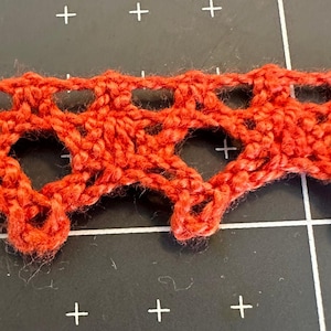 May include: Close-up of a section of red crocheted lace trim. The trim features a repeating pattern of open heart shapes and small loops. The yarn appears to be a textured, possibly wool, material.