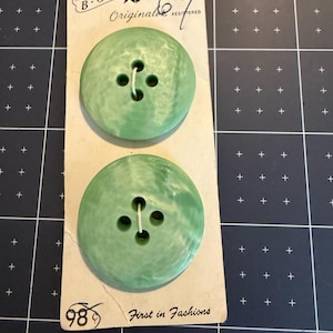 May include: Two round, light green buttons with a marbled texture and four holes each, attached to a rectangular off-white cardboard backing. The card features the text B.G.E. Originals, Washable, and First in Fashions with a crossed-out 98.