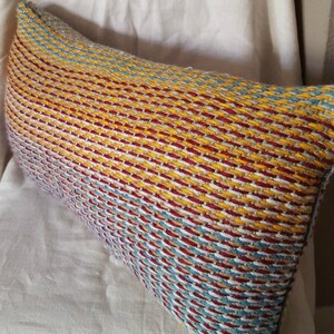May include: A long, rectangular pillow with a woven pattern in shades of red, yellow, blue, and white. The pillow is made of a soft, textured fabric.