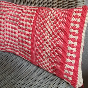 Puede incluir: Un cojín tejido de color rosa y blanco con un patrón geométrico. El cojín está sentado en una silla de mimbre.