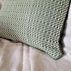 Puede incluir: Una almohada de tiro verde y blanca con textura tejida. La almohada está sobre una colcha de lino blanco.