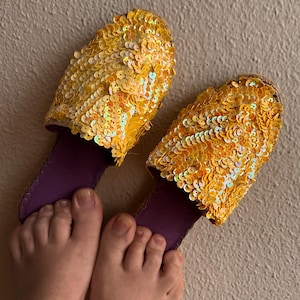 May include: A pair of yellow sequined slippers with purple lining. The slippers are worn on a person's feet.