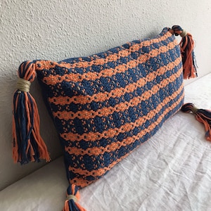 Puede incluir: Una almohada rectangular con un patrón geométrico azul y naranja. La almohada tiene borlas en cada extremo.