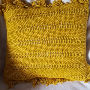 Puede incluir: Una almohada amarilla con textura tejida y ribete de flecos. La almohada está sobre una superficie blanca.