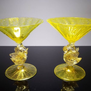 May include: Two yellow glass martini glasses with swan-shaped bases. The glasses are clear with a textured surface and have a gold-colored finish.