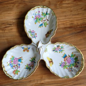 May include: A white porcelain three-section serving dish shaped like three conjoined scallop shells. The dish features hand-painted floral motifs in pink, blue, and green, along with colorful butterfly illustrations and a gold-trimmed scalloped edge.