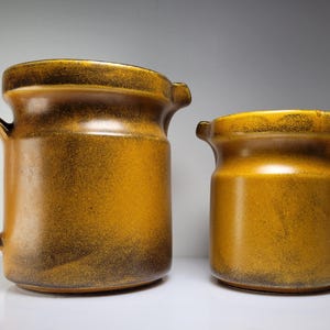 May include: Two ceramic pitchers with a warm, earthy color palette. The larger pitcher has a curved handle and a spout, while the smaller one shares a similar design. Both have a speckled, textured finish.