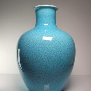 May include: A turquoise ceramic vase with a crackle glaze finish. The vase has a rounded body and a long, slender neck. The rim is a lighter shade of blue. The vase is approximately 10 inches tall.