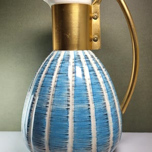 May include: A thermal carafe featuring a white and blue vertically striped pattern with a brushed gold metal collar and a matching curved gold handle. The top section includes a white spout and lid assembly secured with two visible metal rivets.