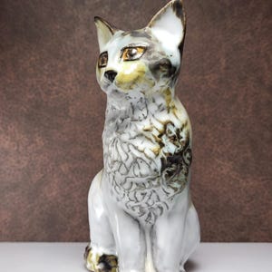 May include: A ceramic cat figurine with a white, gray, and brown mottled glaze. The cat is sitting with its head tilted to the side and its eyes are open.