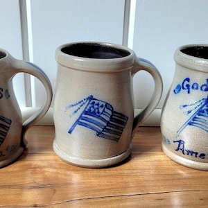 May include: Three beige stoneware mugs with a dark interior and a handle on the right side. Two mugs feature a blue hand-painted American flag and the text God Bless America. The center mug features only a blue hand-painted American flag design.