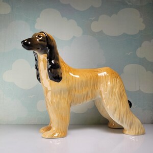 May include: A ceramic Afghan Hound figurine in a standing pose. The dog has a tan body with long, flowing fur, a black face, and black ear accents. The figurine is glossy and detailed, with a classic, elegant design.